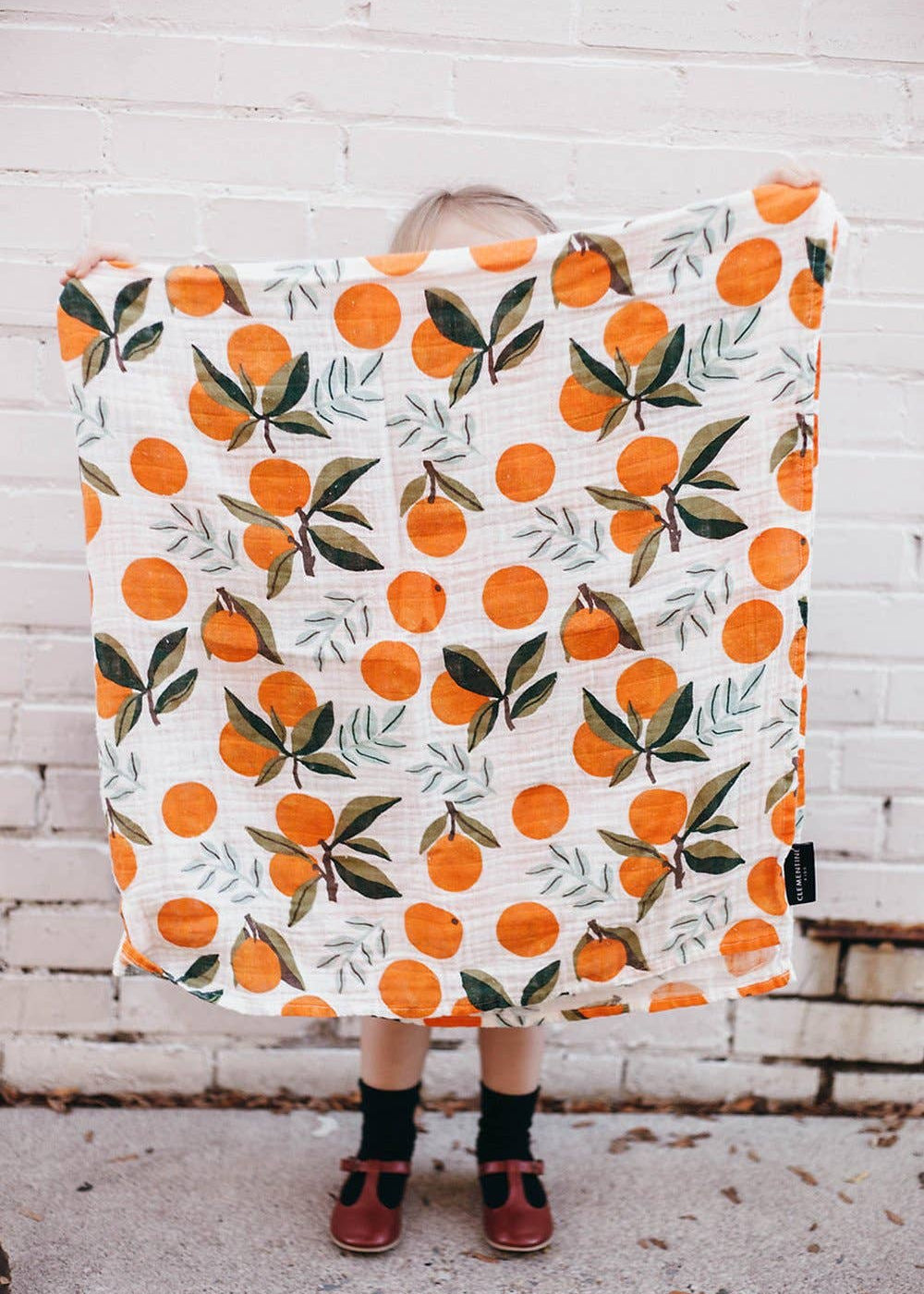 Person holding a blanket with orange pattern against a white brick wall.