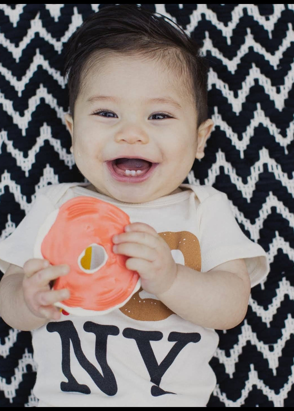 A smiling baby wearing a white short-sleeve t-shirt with 'NY' printed on the front, holding a bright orange circular toy, set against a black and white chevron patterned blanket.