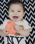 A smiling baby wearing a white short-sleeve t-shirt with 'NY' printed on the front, holding a bright orange circular toy, set against a black and white chevron patterned blanket.