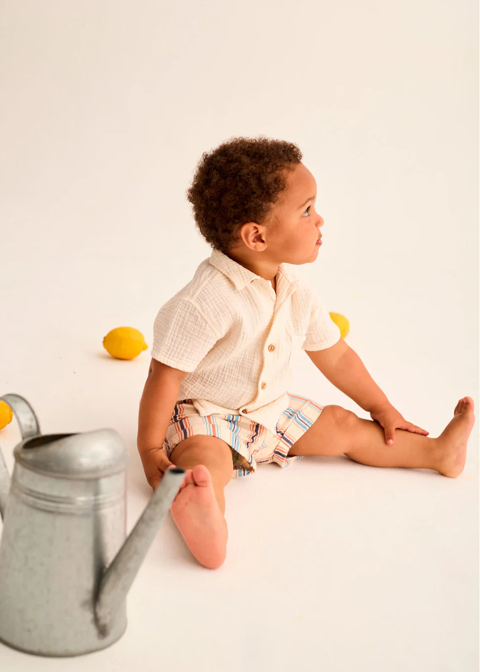 Child sitting on a white surface with lemons and a watering can, wearing a light-colored shirt and shorts.