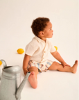 Child sitting on a white surface with lemons and a watering can, wearing a light-colored shirt and shorts.
