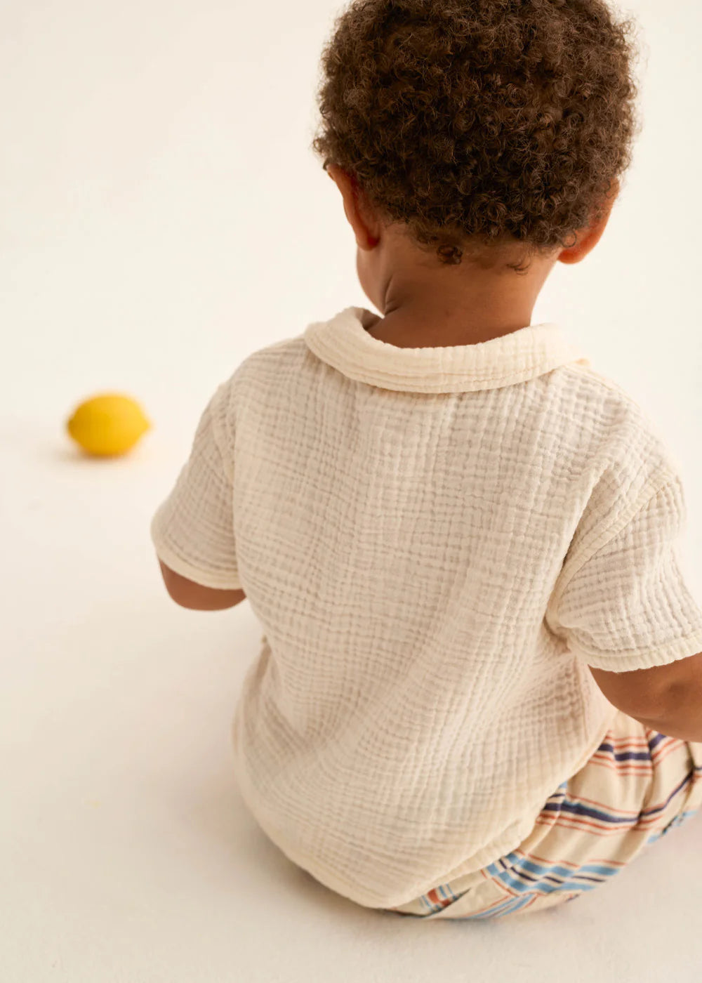 Child sitting on a light surface wearing a textured white shirt and plaid pants.