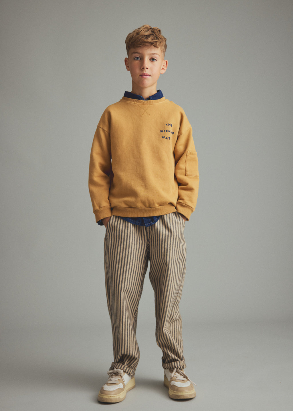 A young boy stands against a neutral gray studio background wearing a mustard yellow sweatshirt with a navy blue collar, beige and navy vertically striped trousers, and beige sneakers.