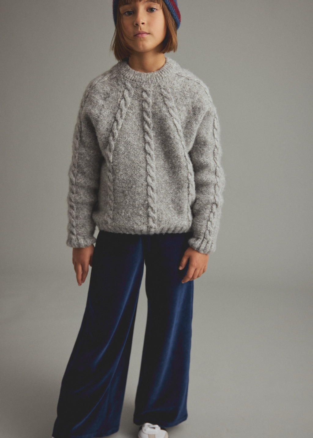 A child model wearing a light gray cable-knit sweater, dark blue wide-leg velvet pants, and a colorful beanie, standing against a neutral gray studio background.