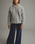 A child model wearing a light gray cable-knit sweater, dark blue wide-leg velvet pants, and a colorful beanie, standing against a neutral gray studio background.