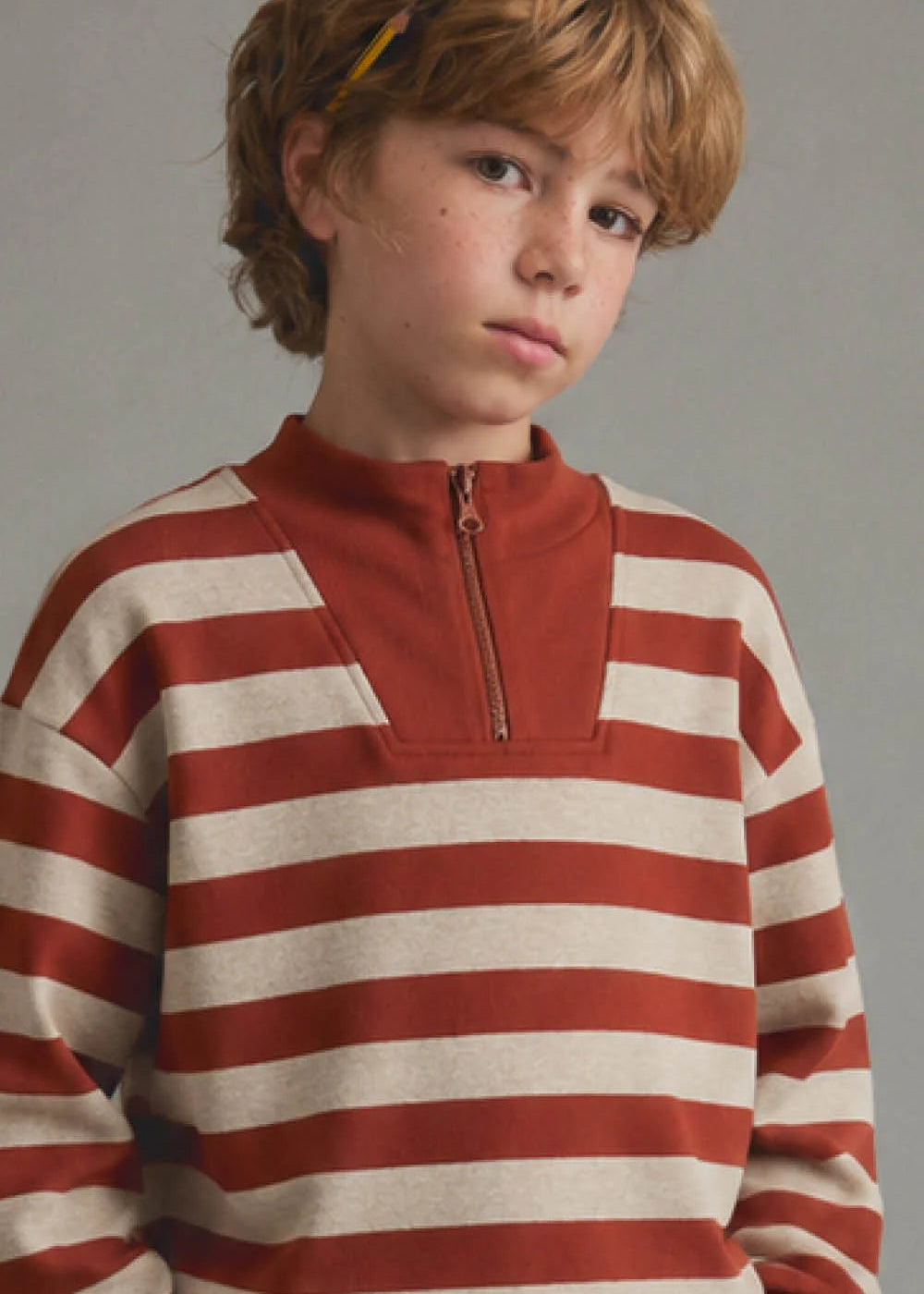 A young boy with light brown hair wearing a rust red and cream striped quarter-zip sweatshirt, photographed against a neutral gray studio background.
