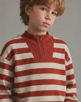 A young boy with light brown hair wearing a rust red and cream striped quarter-zip sweatshirt, photographed against a neutral gray studio background.