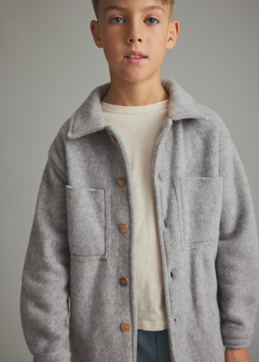 A young boy wearing a light gray wool-blend button-up jacket over a cream t-shirt, photographed against a solid gray studio background.
