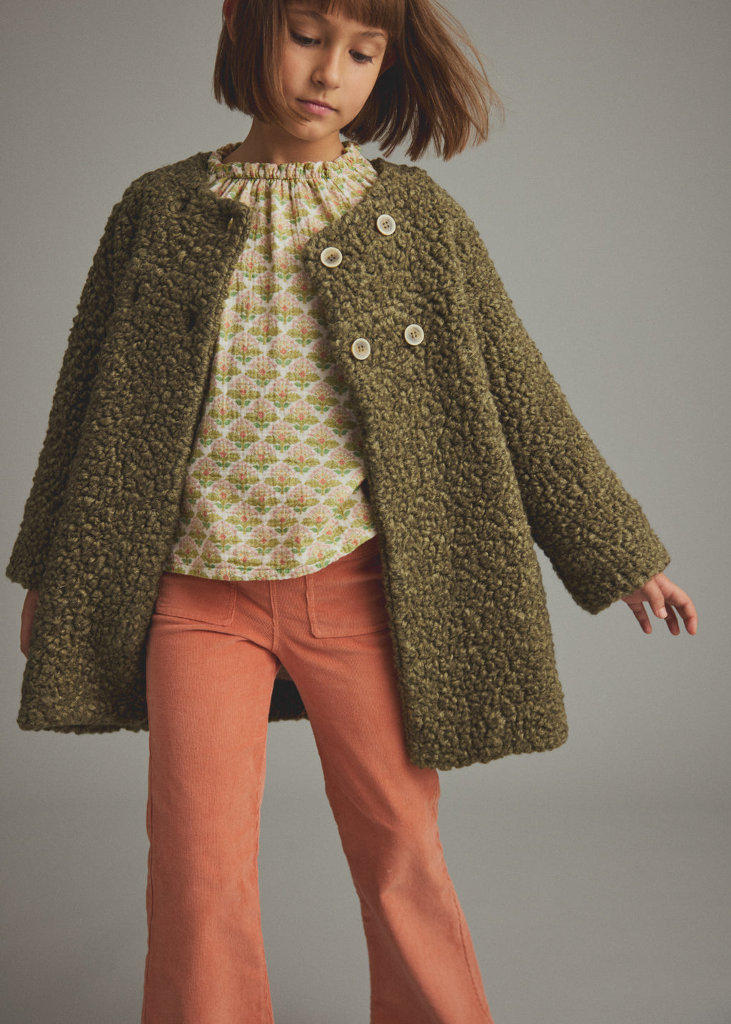 A child model wearing a textured olive green boucle-style cardigan over a patterned cream top and rust-orange pants, photographed against a plain gray studio background.