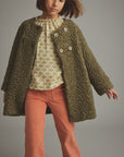 A child model wearing a textured olive green boucle-style cardigan over a patterned cream top and rust-orange pants, photographed against a plain gray studio background.