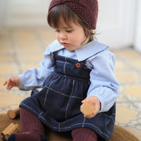 Child wearing a blue dress with plaid pattern and maroon headband and tights, sitting on a wooden floor.