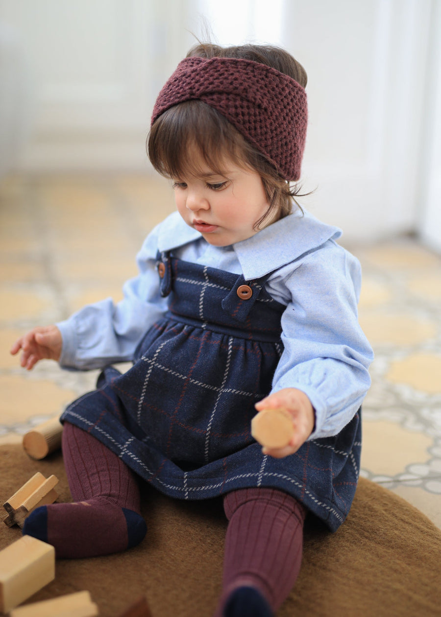 Child wearing a blue dress with plaid pattern and maroon headband and tights, sitting on a wooden floor.