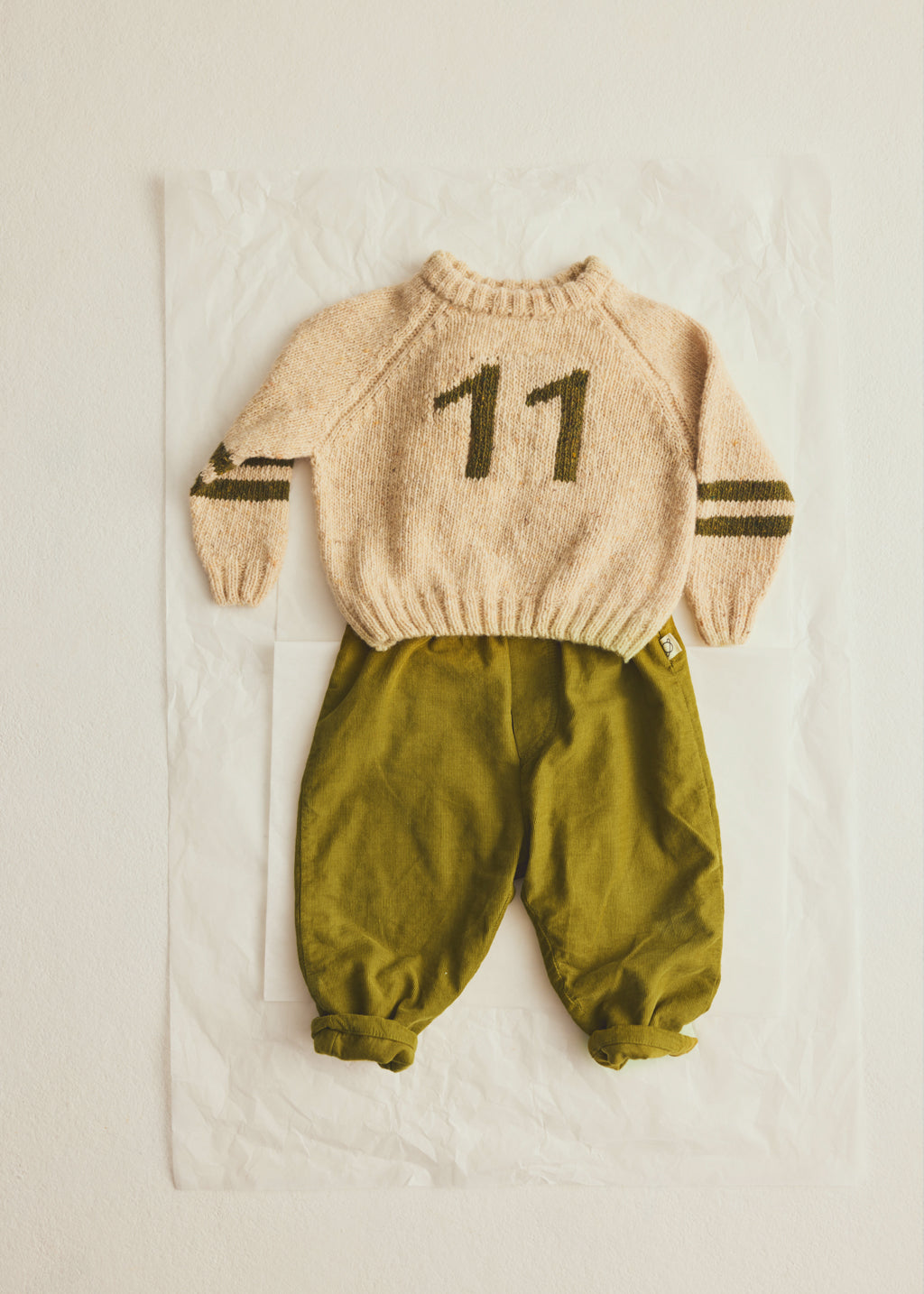 A baby knit two-piece outfit featuring a cream-colored long-sleeve sweater with the number '11' and dark olive stripes on the sleeves, paired with olive green pants, laid flat on a white sheet.