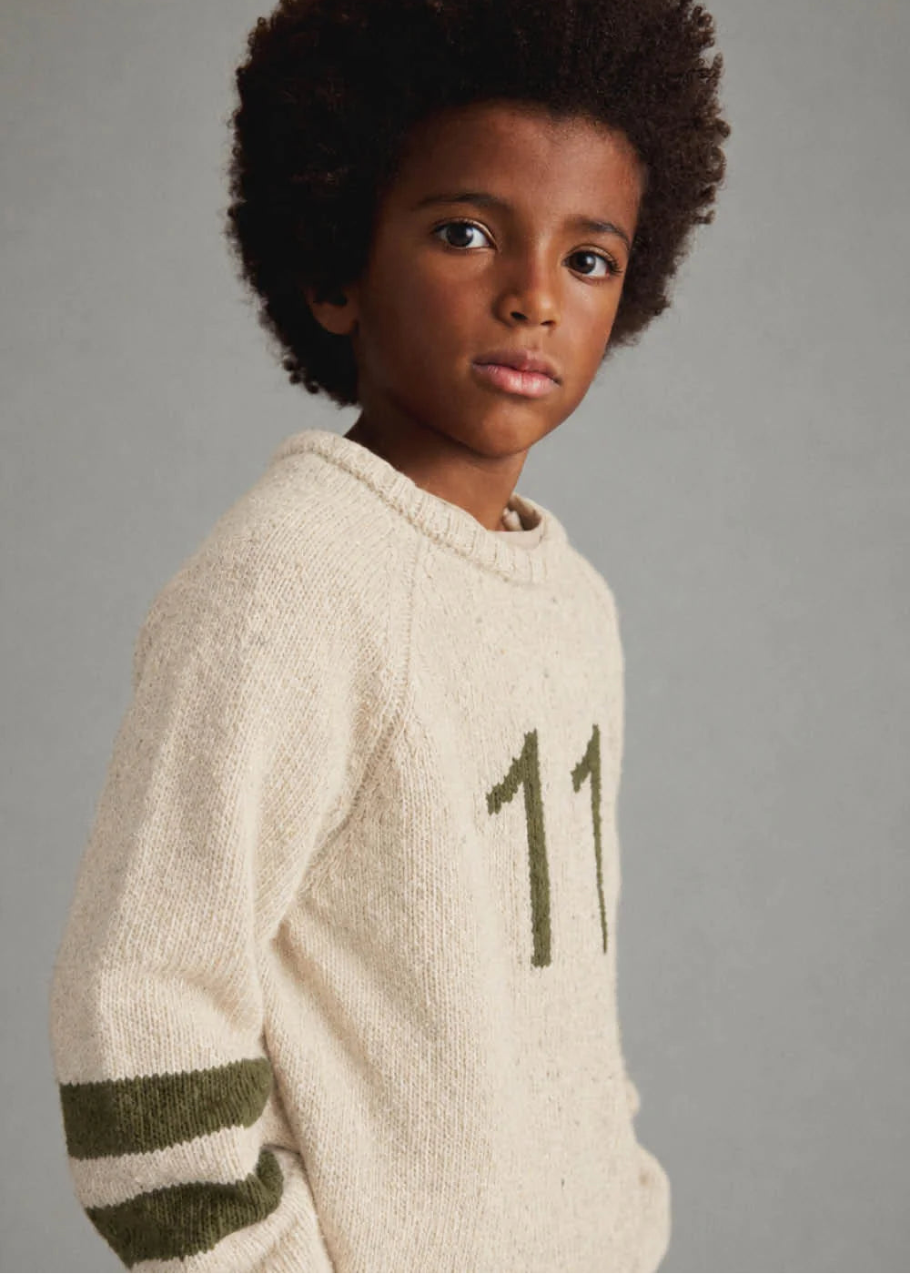 A child model wearing a cream-colored knit sweater with olive green stripes on the sleeves and a large olive green number '11' on the chest, set against a plain gray studio background.