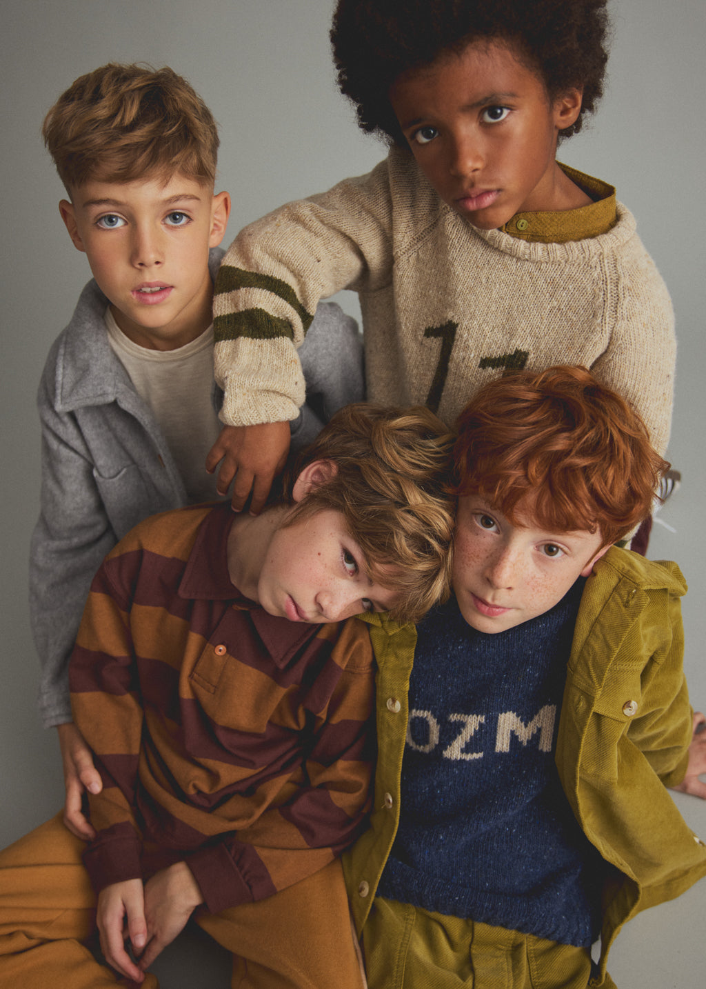 Four children posing together in cozy, vintage-style knit and cotton clothing against a solid gray studio background.