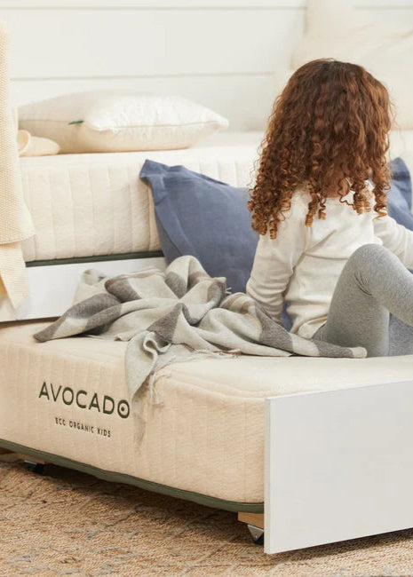Child sitting on a bed with 'Avocado' branding, wearing a white sweater and gray leggings.