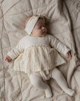 Baby in a white outfit with a textured skirt lying on a beige blanket.