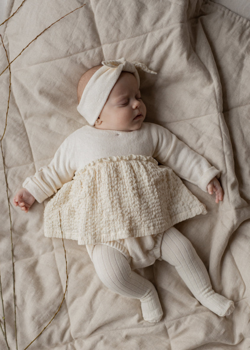 Baby in a white outfit with a textured skirt lying on a beige blanket.