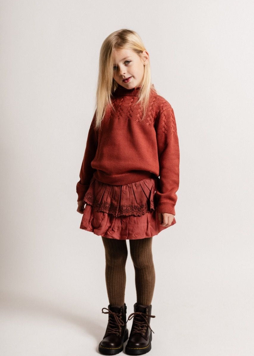 Young girl wearing a red sweater and skirt with brown tights and shoes on a white background