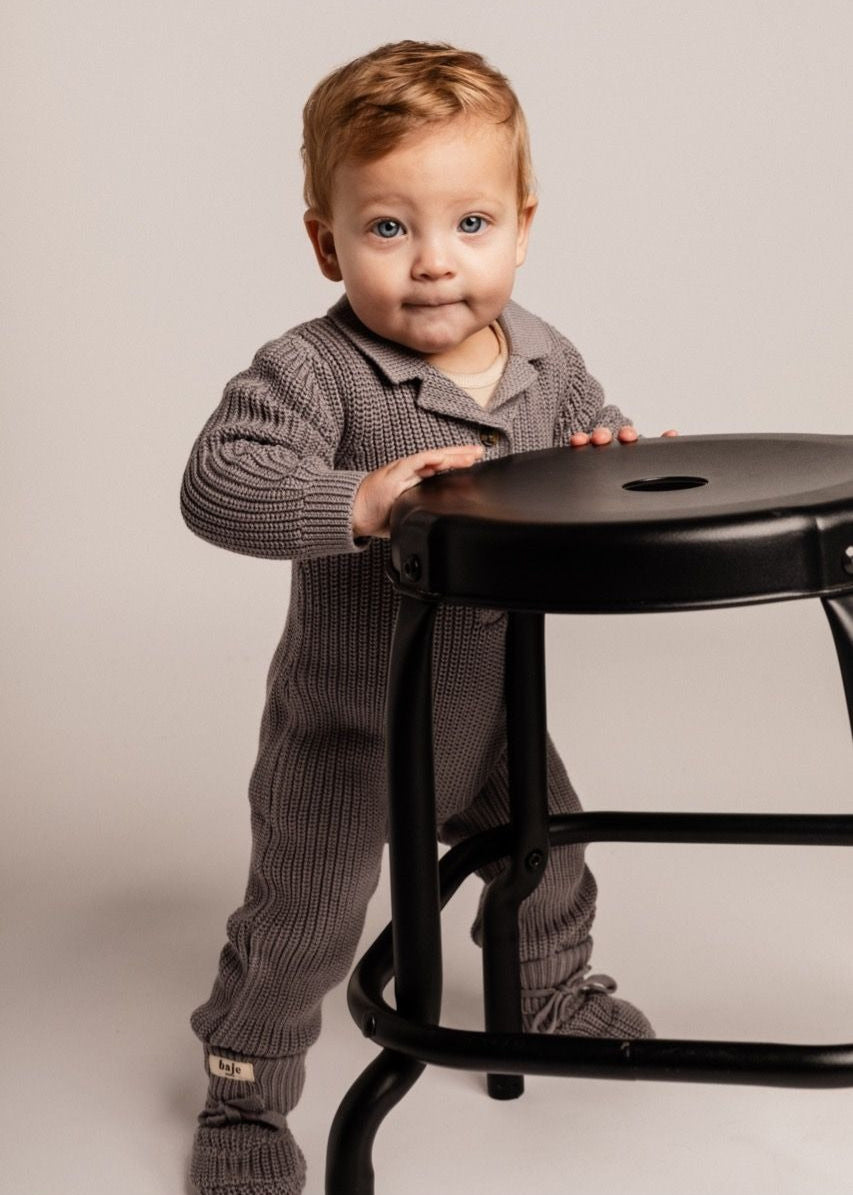 Child in a gray outfit standing next to a black stool on a plain background