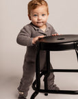 Child in a gray outfit standing next to a black stool on a plain background