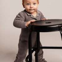 Child in a gray outfit standing next to a black stool on a plain background