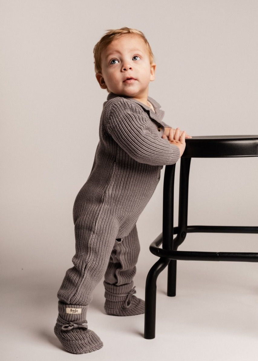 Child wearing a gray ribbed outfit standing next to a black chair on a plain background
