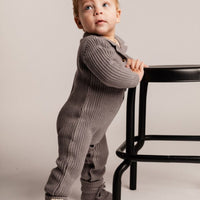 Child wearing a gray ribbed outfit standing next to a black chair on a plain background