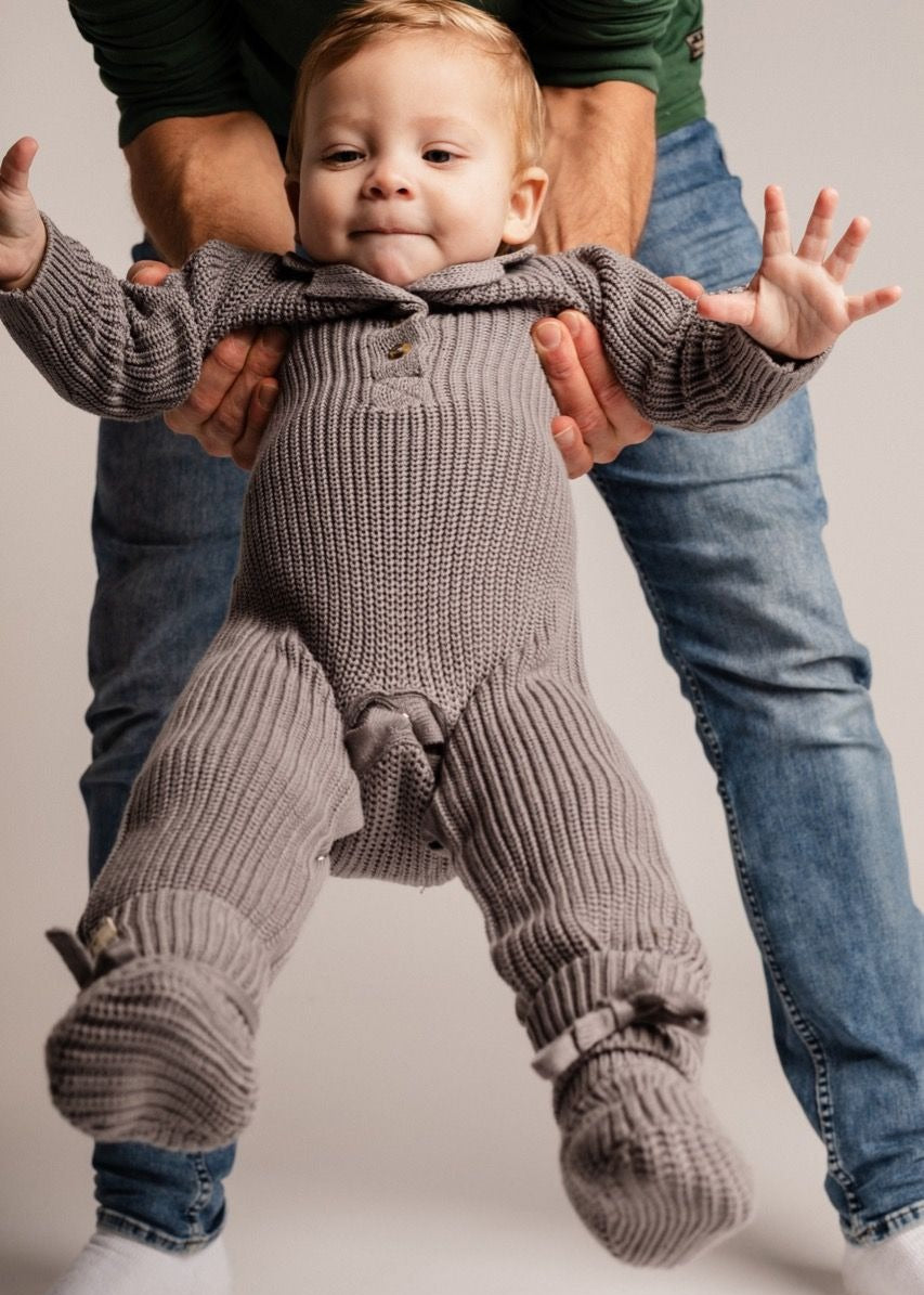 Baby in a knitted outfit being held up by an adult on a plain background