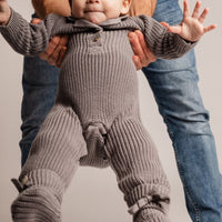 Baby in a knitted outfit being held up by an adult on a plain background