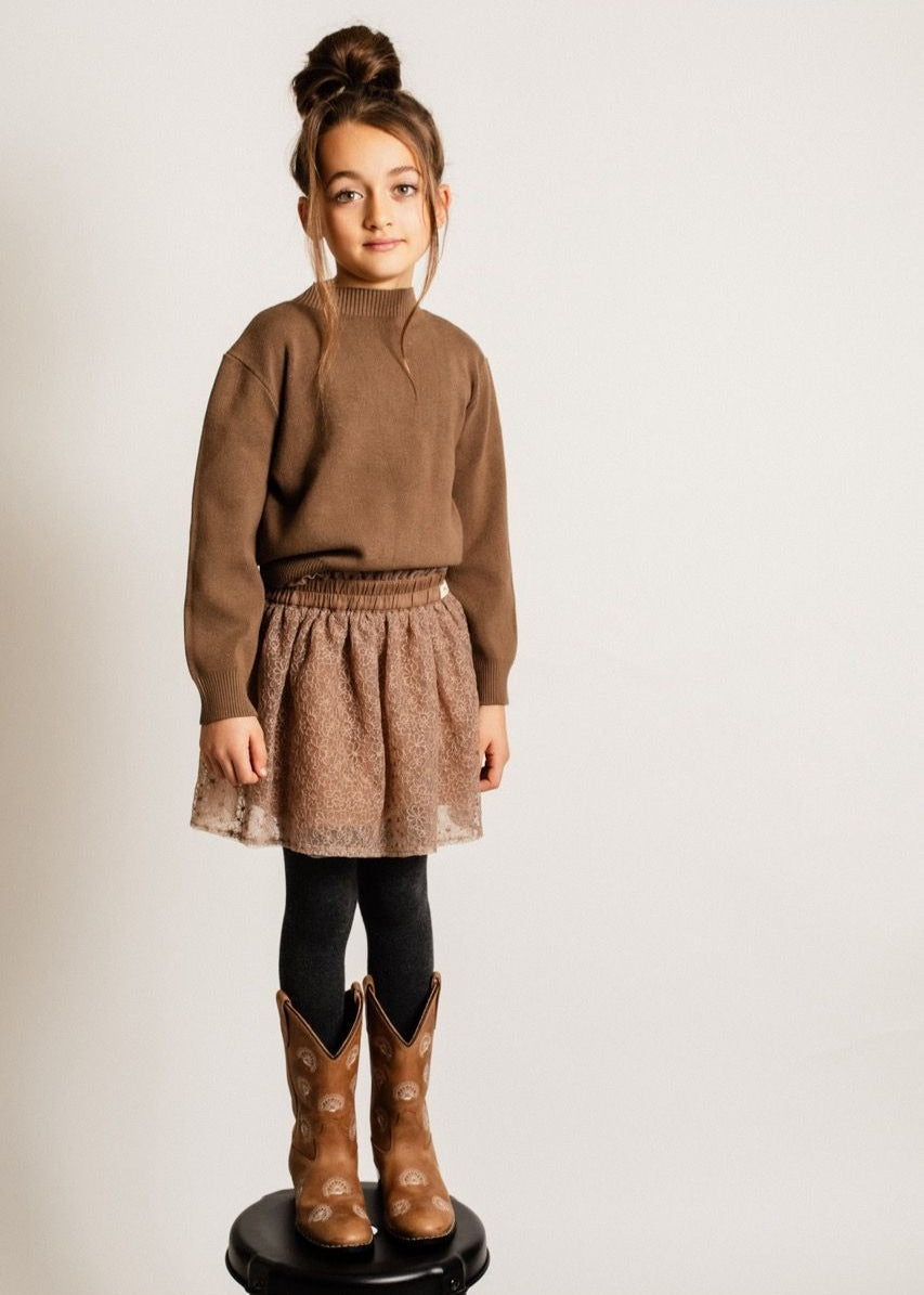 Child wearing a brown sweater, lace skirt, and cowboy boots on a plain background