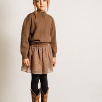 Child wearing a brown sweater, lace skirt, and cowboy boots on a plain background