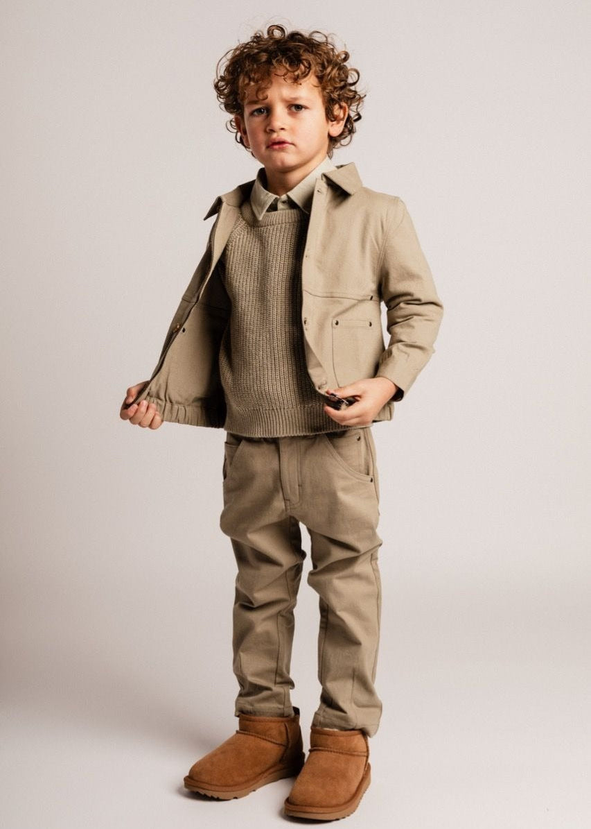 Child wearing a beige coat, sweater, pants, and boots on a plain background