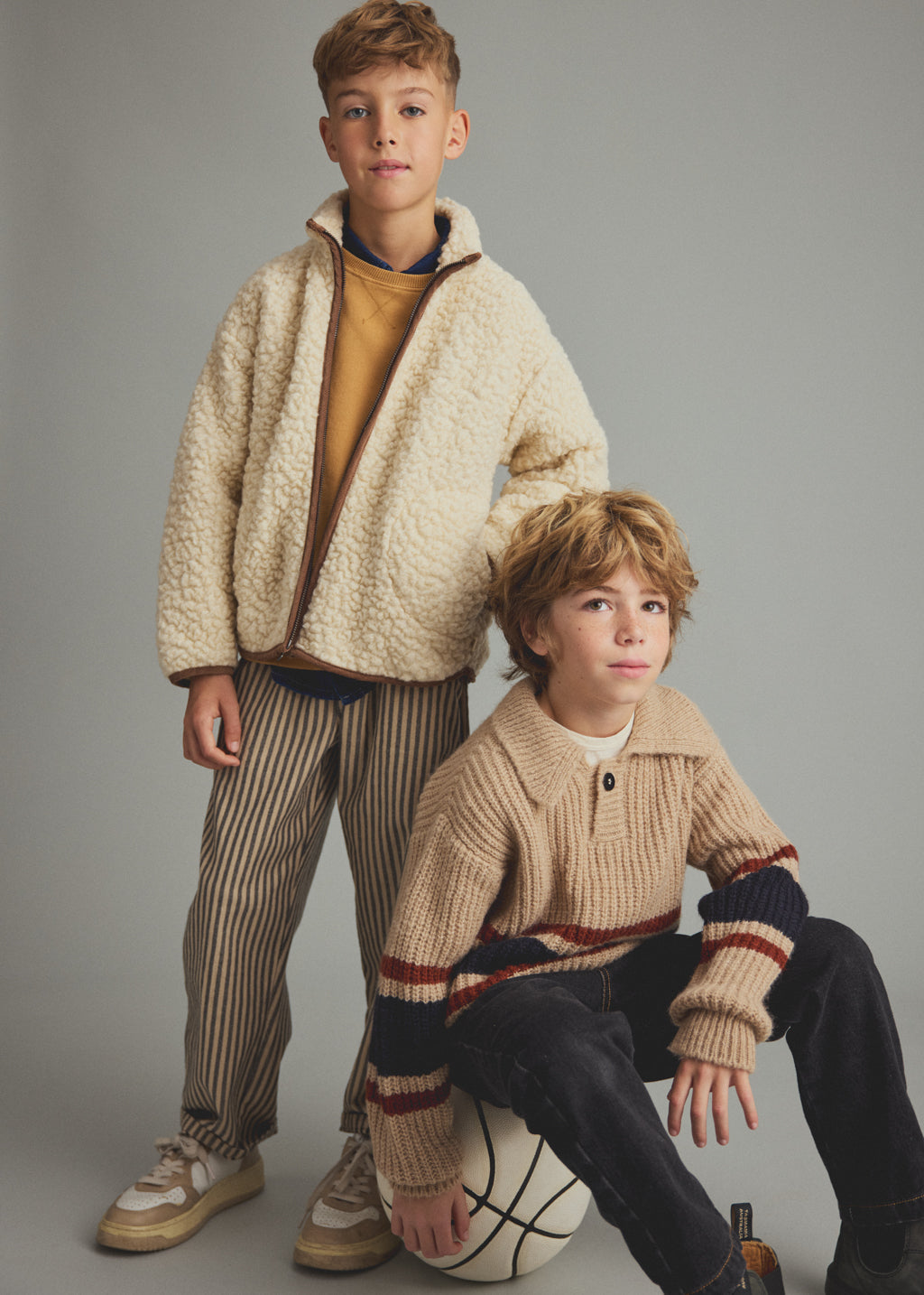 Two boys modeling casual knit and fleece clothing against a solid gray studio background. The standing boy wears a cream sherpa zip-up jacket over a mustard yellow sweater and brown-and-beige striped pants.