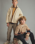 Two boys modeling casual knit and fleece clothing against a solid gray studio background. The standing boy wears a cream sherpa zip-up jacket over a mustard yellow sweater and brown-and-beige striped pants.