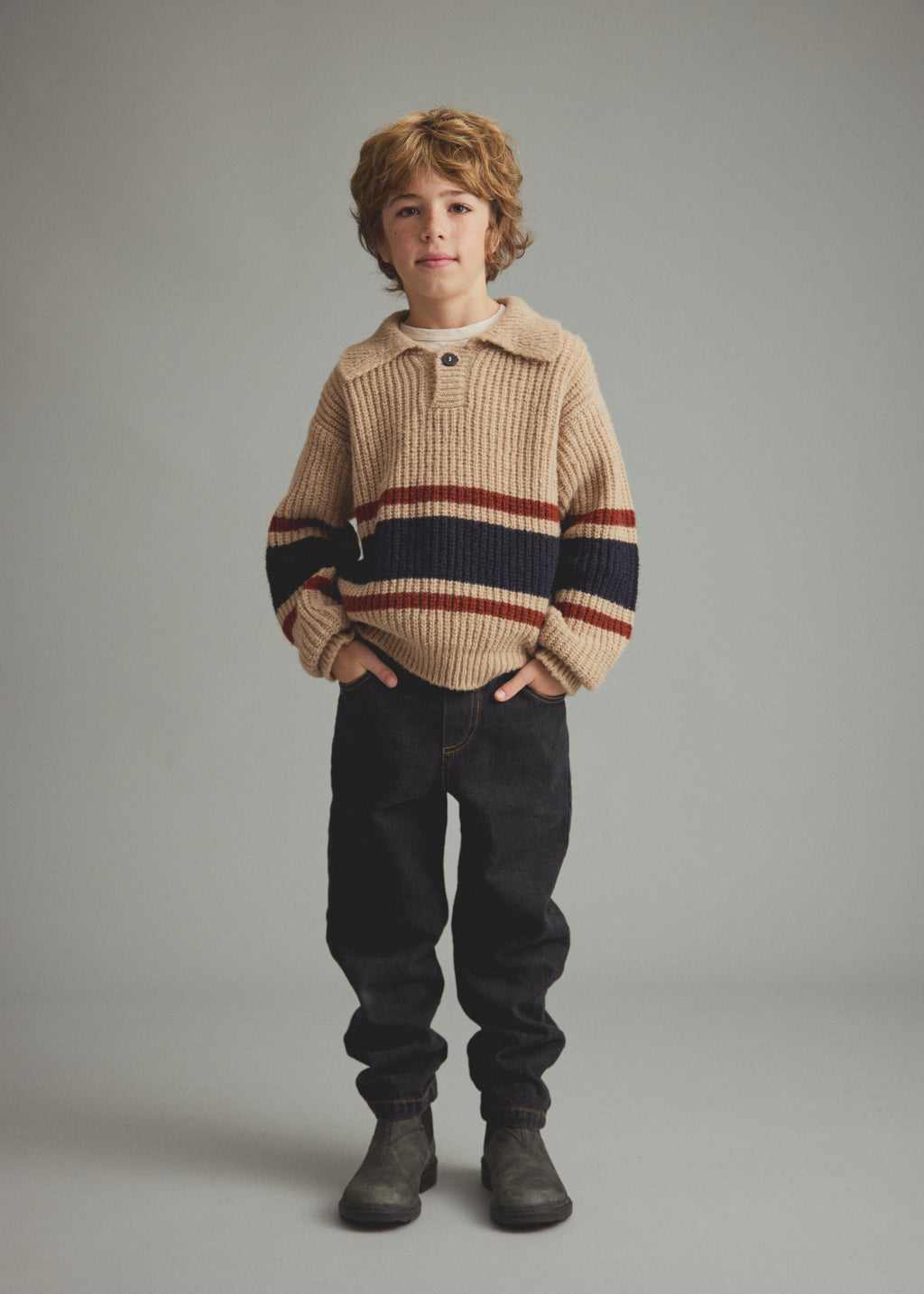 A young boy stands against a neutral gray studio background wearing a beige ribbed knit sweater with horizontal navy and rust-red stripes, dark pants, and dark boots.