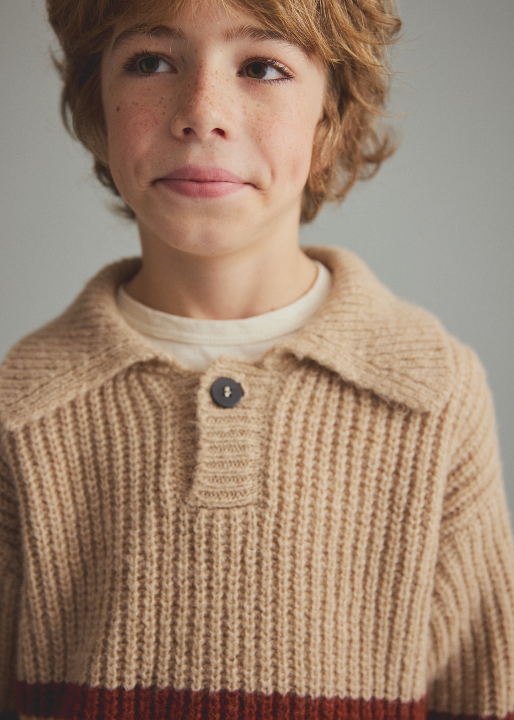 A close-up of a child wearing a beige ribbed knit polo cardigan with a dark button, layered over a cream shirt, against a neutral gray background.