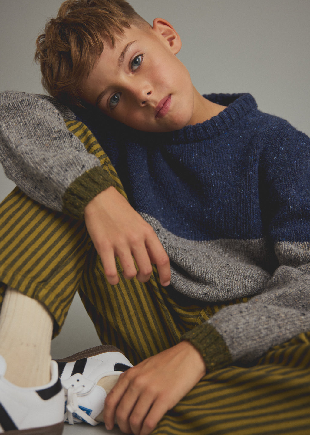 A young boy wearing a navy and gray color-blocked knit sweater, olive green and yellow striped trousers, and white sneakers, posing against a neutral gray background.