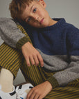 A young boy wearing a navy and gray color-blocked knit sweater, olive green and yellow striped trousers, and white sneakers, posing against a neutral gray background.