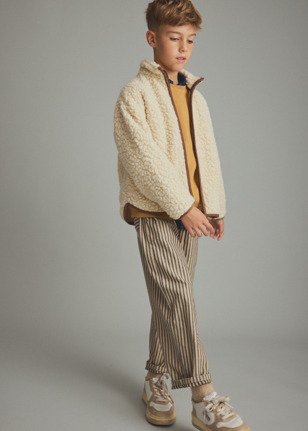 A child model wearing a cream sherpa fleece zip-up jacket over a mustard yellow shirt, paired with beige and brown vertically striped trousers and beige sneakers, set against a plain gray studio background.