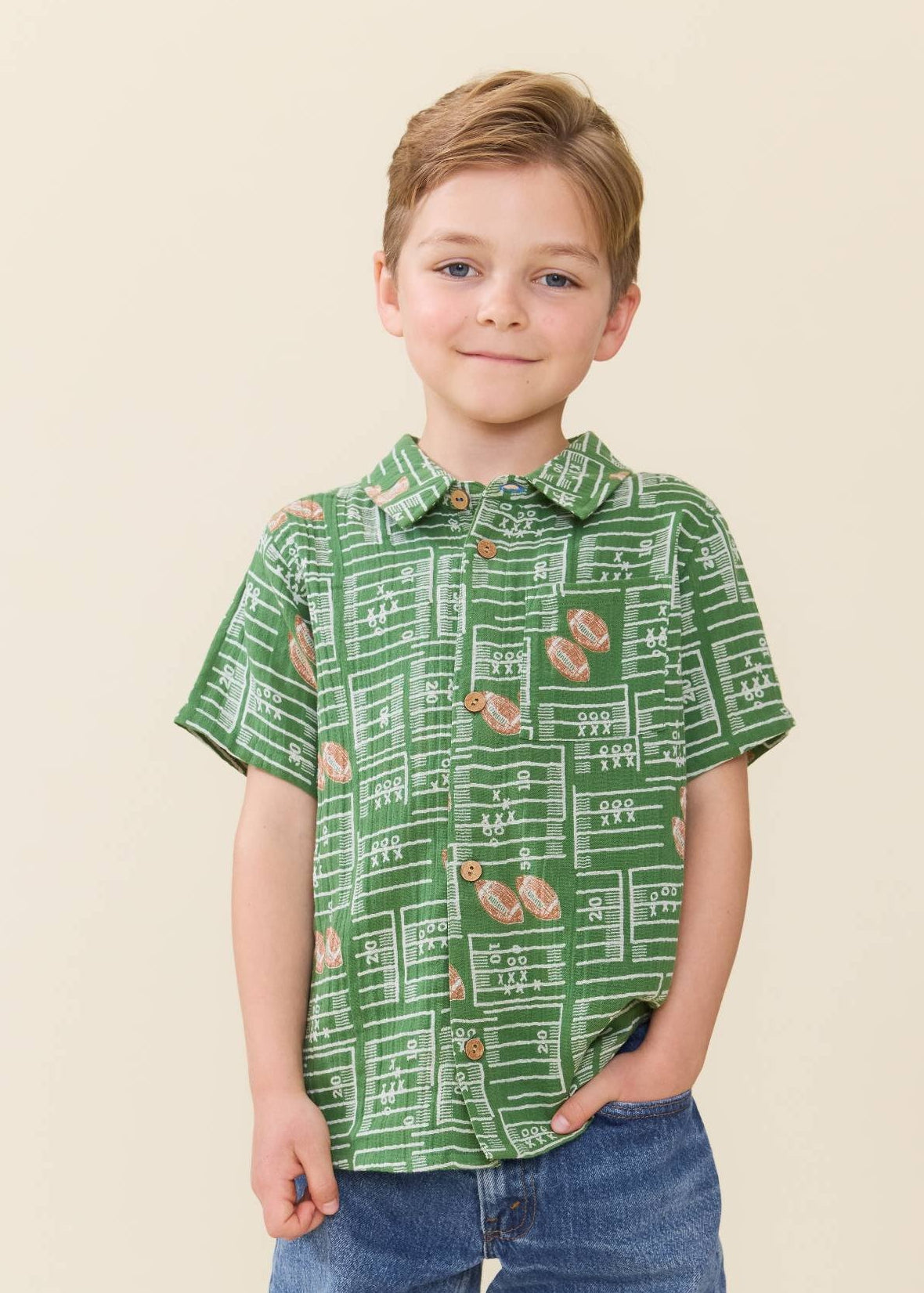 A young boy stands wearing a green short-sleeve button-up shirt with a chalkboard and football pattern, paired with blue jeans, against a warm beige background.