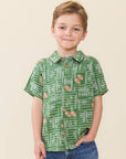 A young boy stands wearing a green short-sleeve button-up shirt with a chalkboard and football pattern, paired with blue jeans, against a warm beige background.
