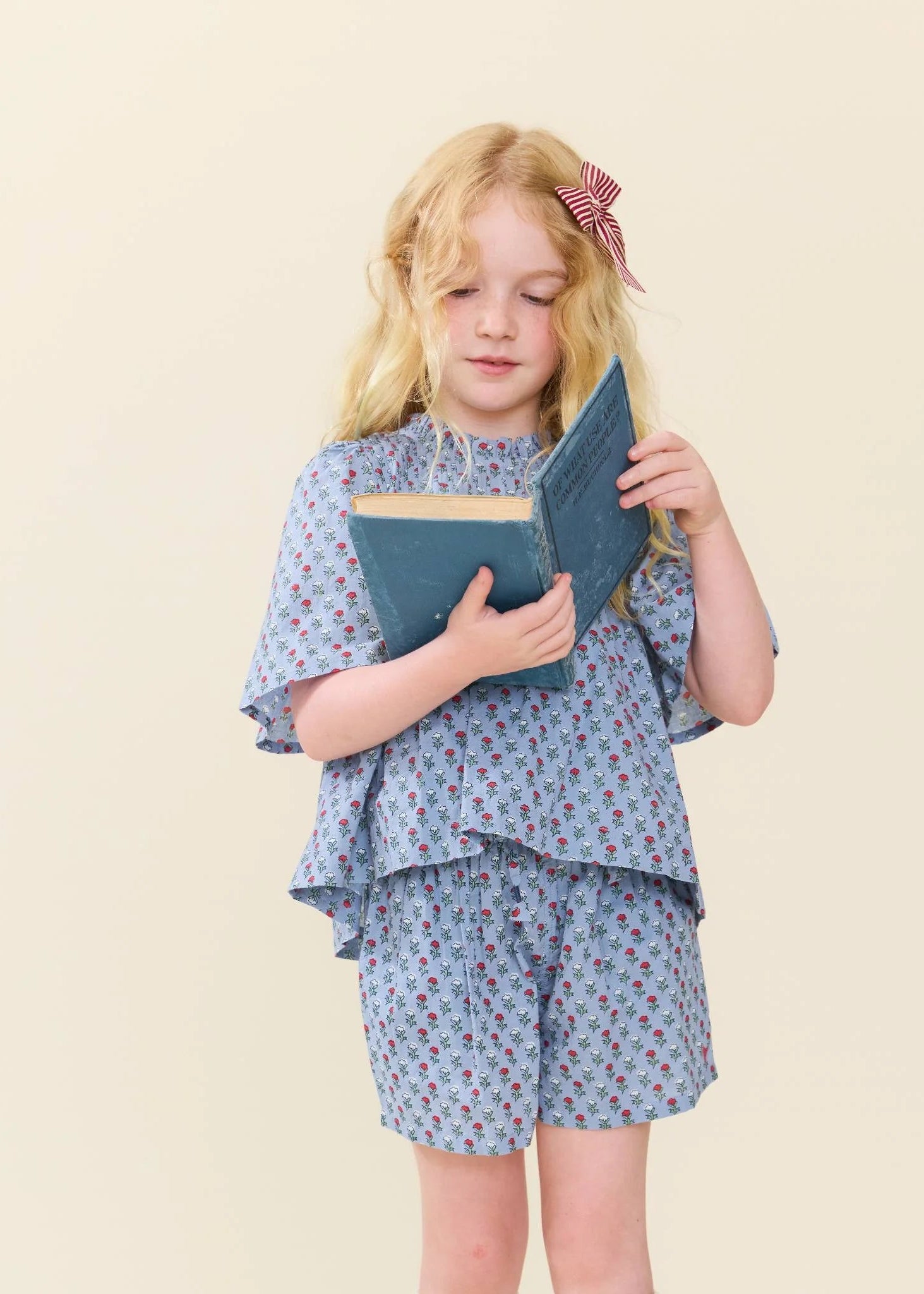 A young girl with blonde hair and a red-and-white bow stands against a plain beige background, holding a blue book. She is wearing a light blue patterned short-sleeve top and matching shorts.