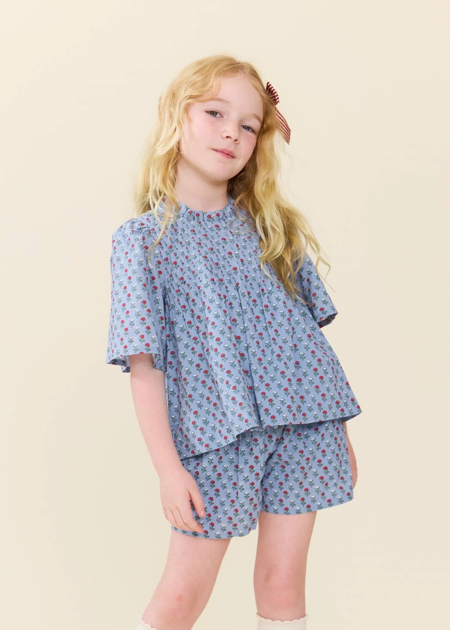 A young girl with blonde hair wearing a light blue short-sleeve top and matching shorts with a small red and pink floral pattern, posed against a solid light beige background.