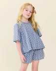 A young girl with blonde hair wearing a light blue short-sleeve top and matching shorts with a small red and pink floral pattern, posed against a solid light beige background.