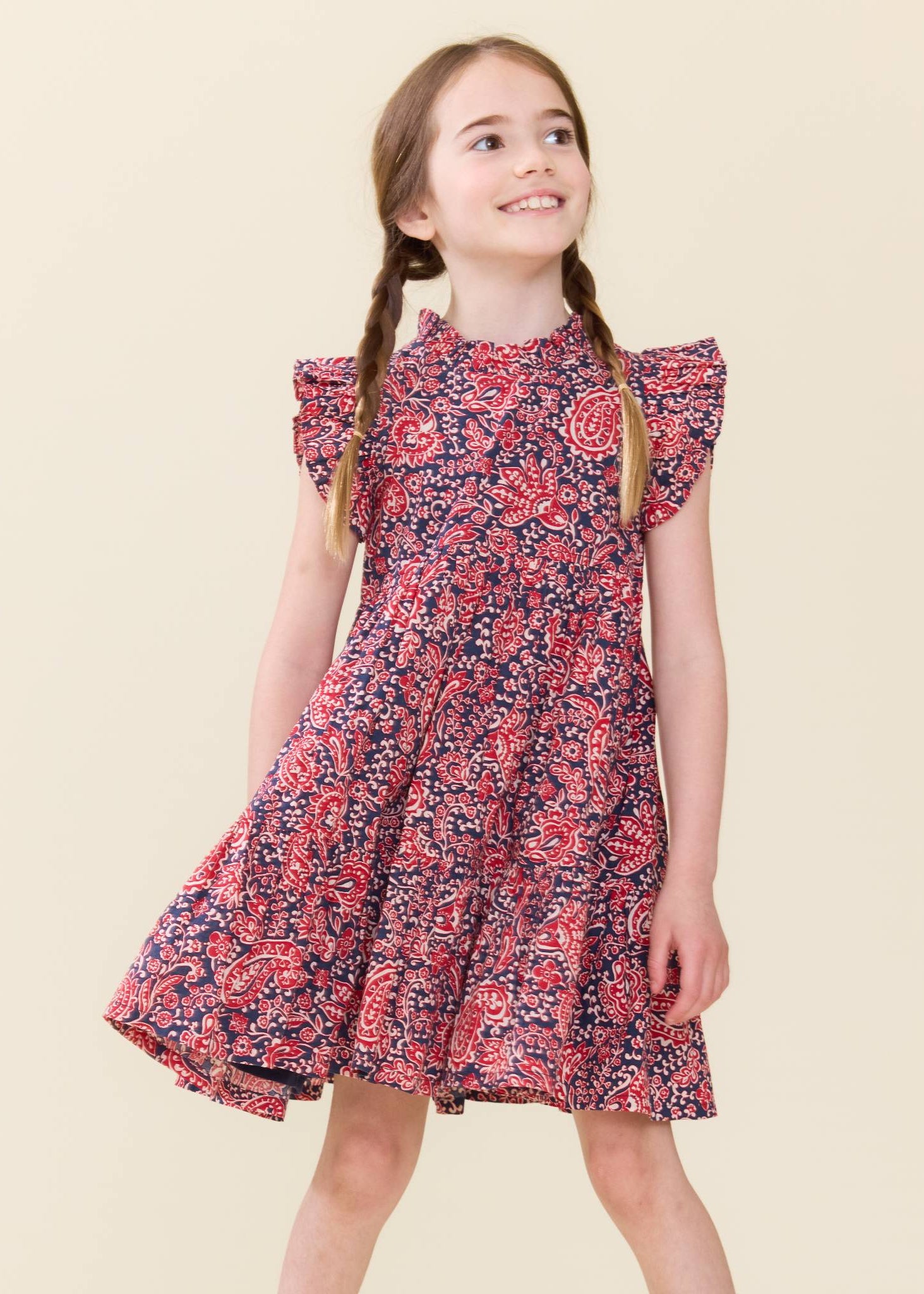 A young girl with braided hair wearing a short-sleeve floral dress with a red, pink, and navy blue pattern, standing against a solid light beige background.