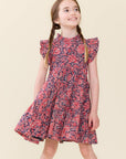 A young girl with braided hair wearing a short-sleeve floral dress with a red, pink, and navy blue pattern, standing against a solid light beige background.