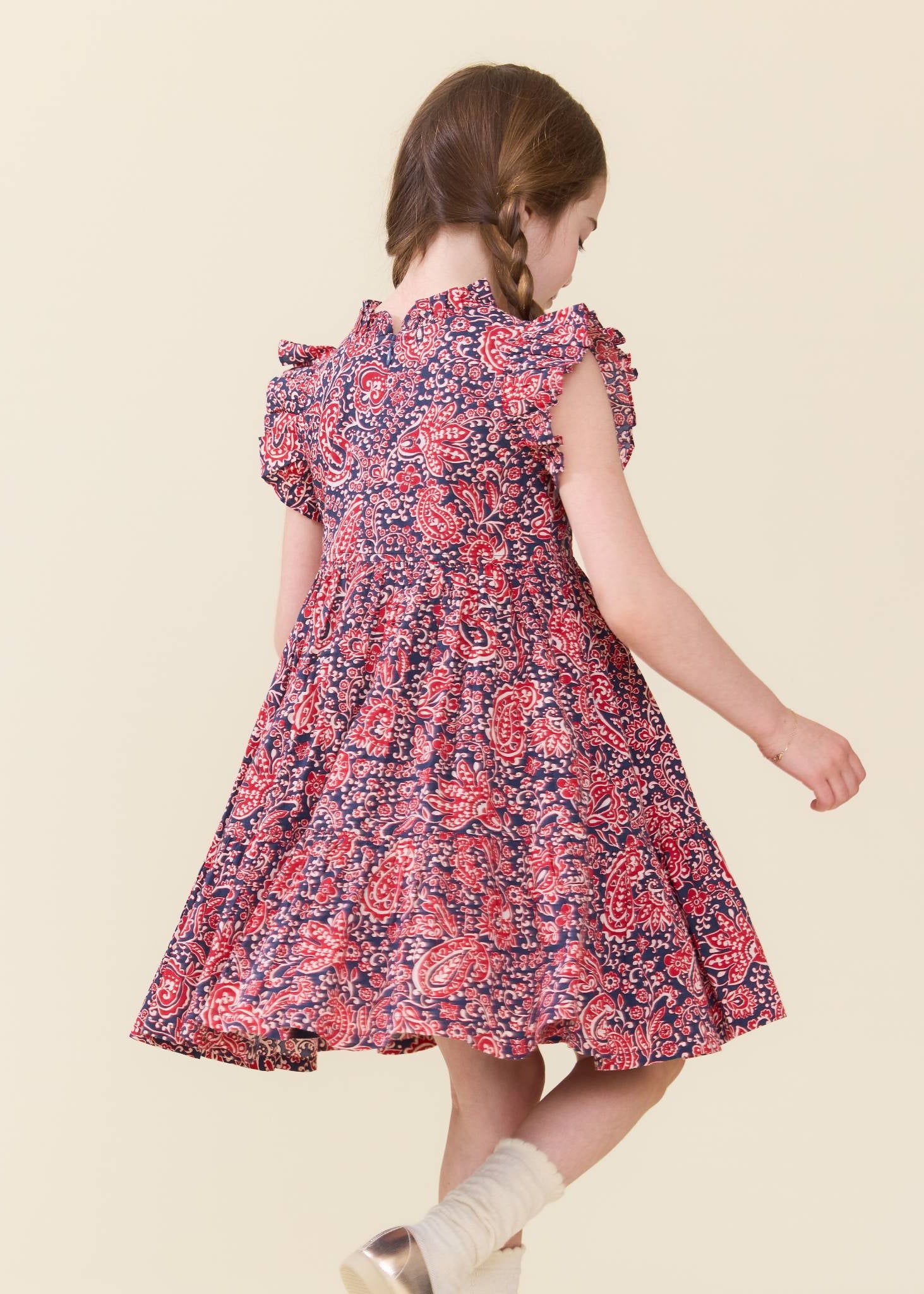 A young girl with braided brown hair is shown from behind wearing a short-sleeved, knee-length floral dress with a red, pink, and navy blue pattern. She is standing against a solid, light beige background.