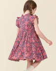 A young girl with braided brown hair is shown from behind wearing a short-sleeved, knee-length floral dress with a red, pink, and navy blue pattern. She is standing against a solid, light beige background.