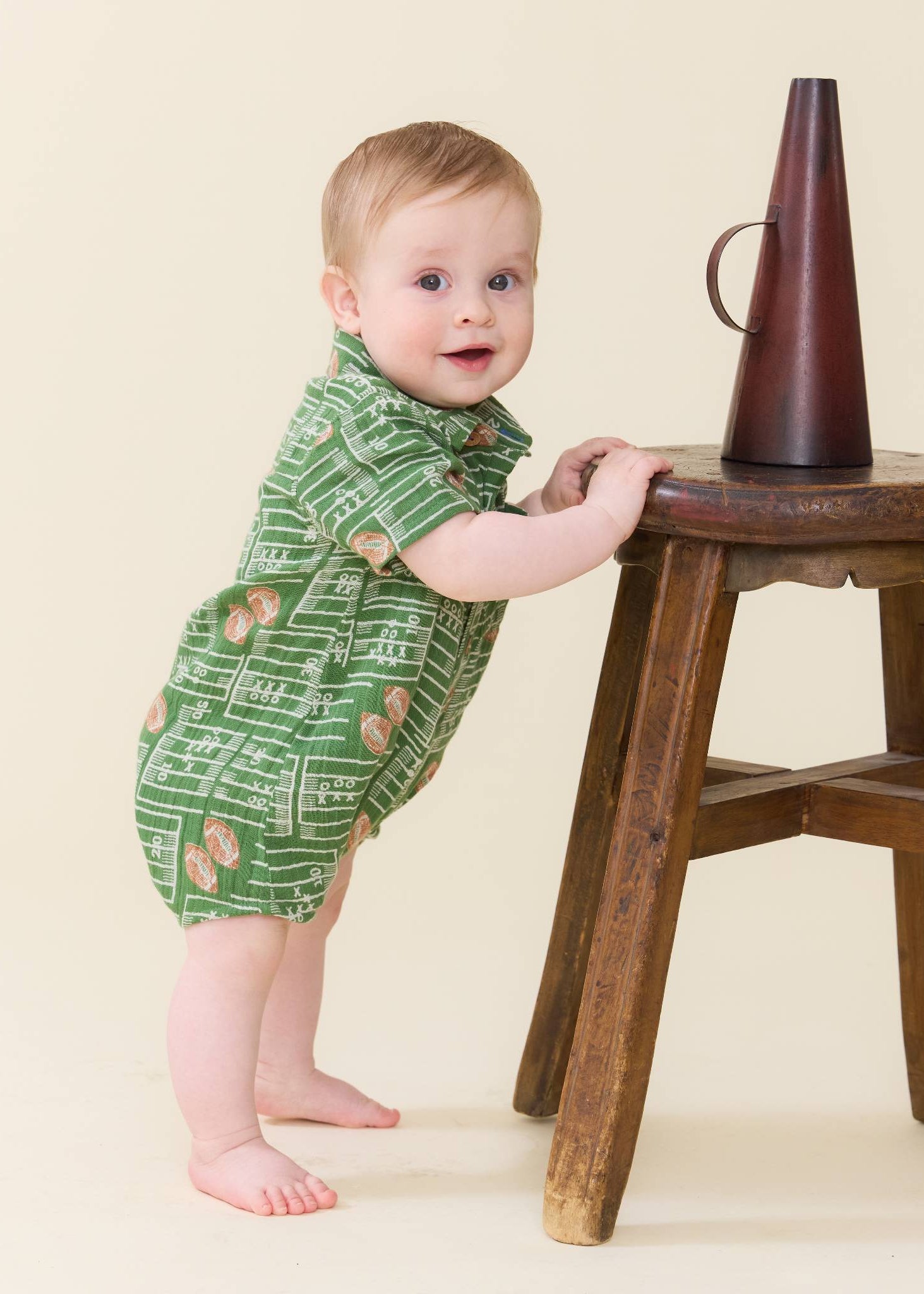 A baby standing on a wooden stool and holding onto it, wearing a green short-sleeve romper with a white musical notation print. A dark brown conical object sits on the stool.